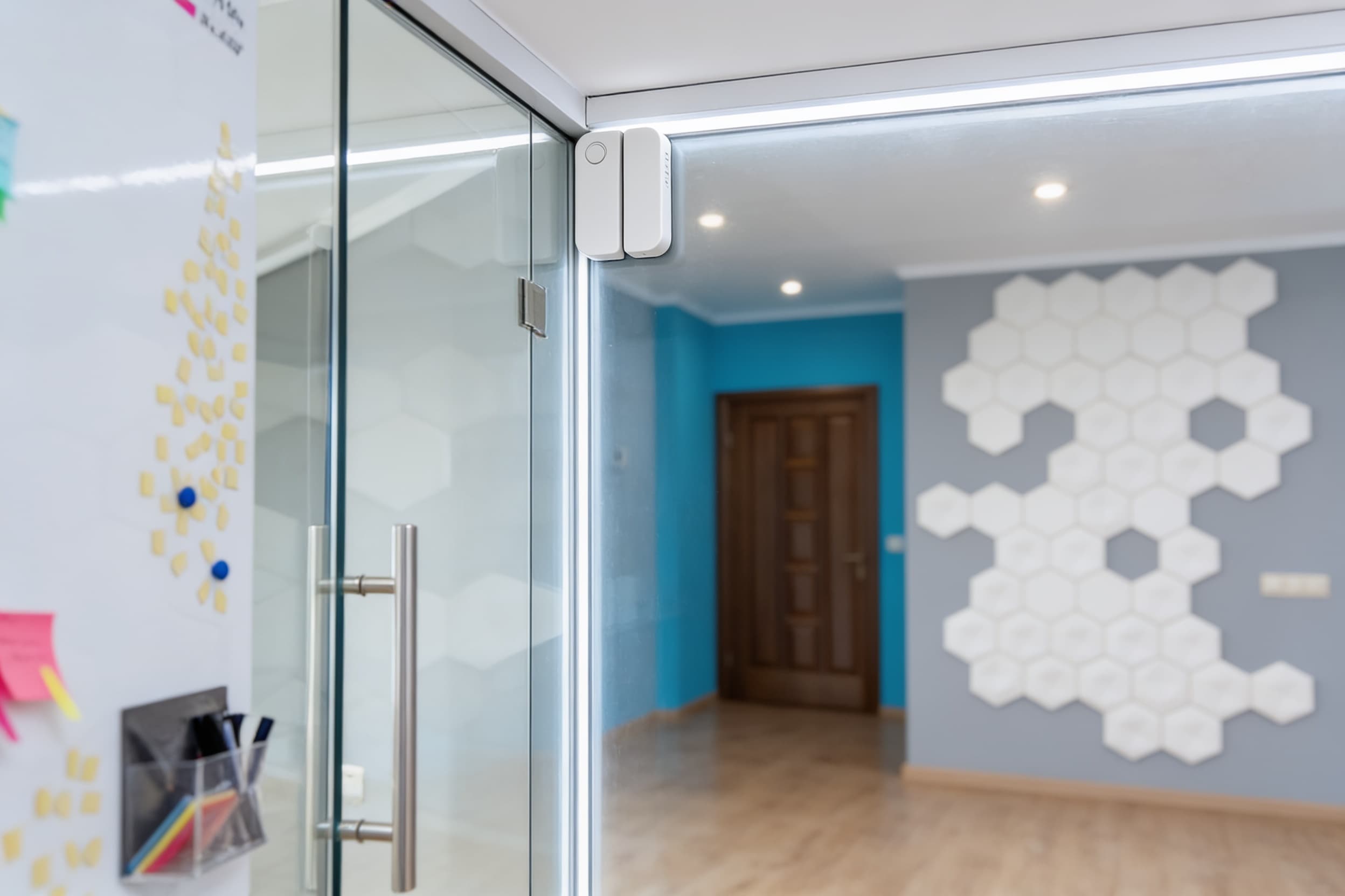 A wall-mounted security sensor both pieces aligned on the glass door edge and frame at the top corner